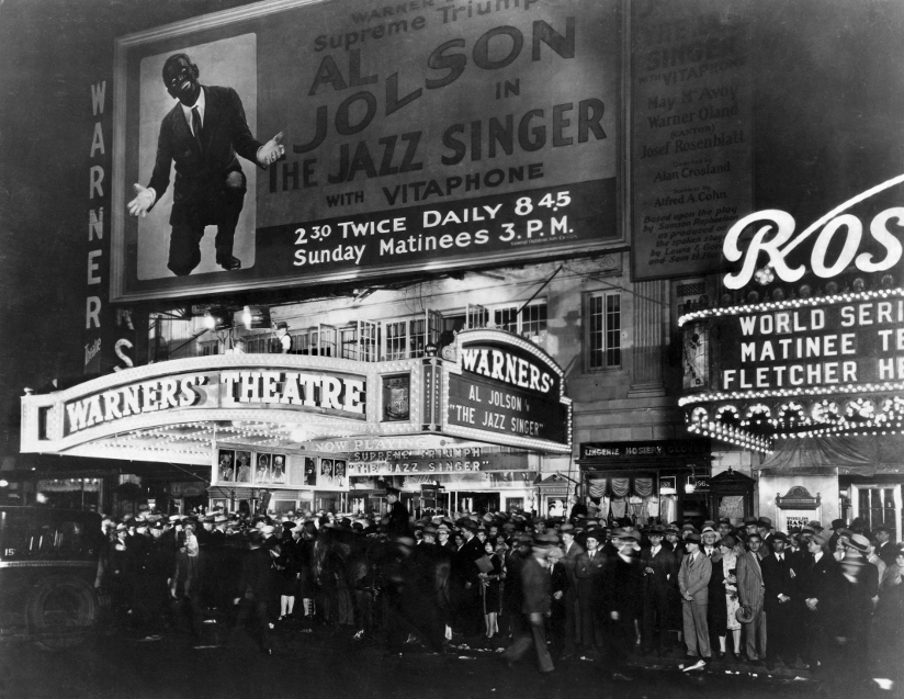 Svart-hvitt fotografi av mennesker som står i kø utenfor Warners’ Theatre for å se <span class="italic">The Jazz Singer</span> i 1927. Over inngangspartiet henger en stor reklametavle (billboard) med filmplakat med opplysninger om tidspunkt for framvisning.