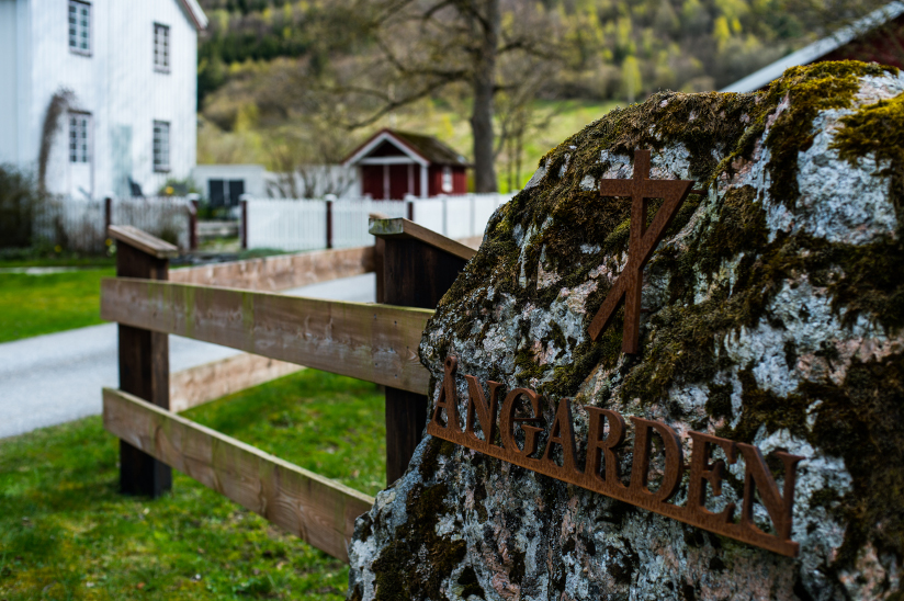 Fotografi med nærbilde av et bumerke som er plassert på en stein. Bumerket følger gården som skimtes i bakgrunn.