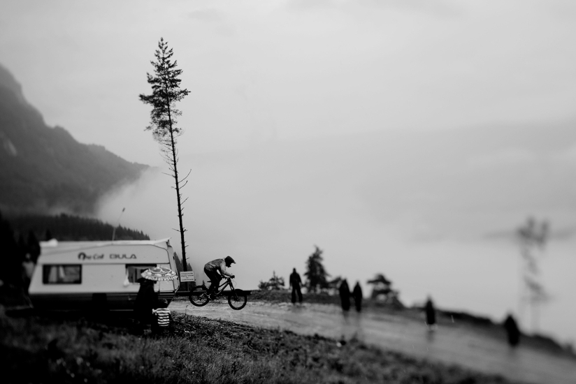 Svart-hvitt fotografi av landskap bestående av fjell og en tåkelagt dal. I forkant ser vi en campingvogn, et høyt furutre, en syklist og mennesker som spaserer i regnet.