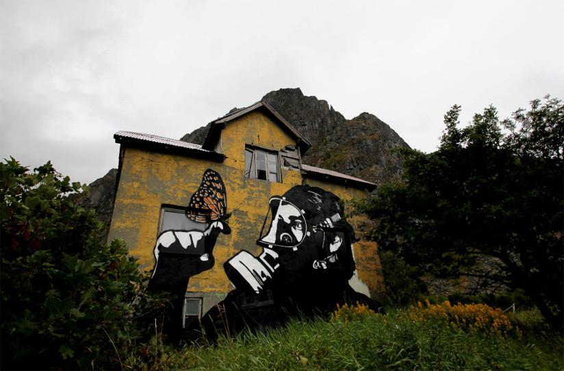 Fotografi av en husvegg med graffiti på veggen. Motivet er nærbilde av en person med gassmaske som ser på en finger der en sommerfugl har landet.