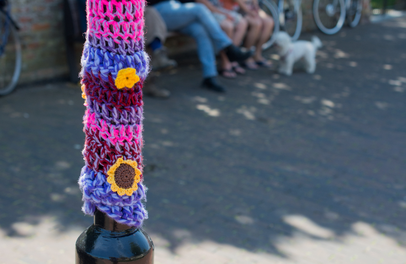 Fotografi av en lyktestolpe i et urbant miljø som er dekket av garn i forskjellige farger (yarnbombing/geriljastrikking). Strikketøyet har et stripete mønster basert på brede masker og er dekortert med to blomster – en gul blomst samt en solskikke.