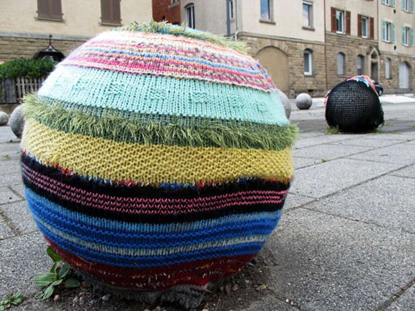 Fotografi av en betongball i et urbant miljø som er dekket av garn i forskjellige farger. (yarnbombing/geriljastrikking). Strikketøyet har stor masker og et stripete mønster. I bakgrunnen skimter vi en tilsvarende ball som er dekket av et stort strikketøy strikket med større masker.