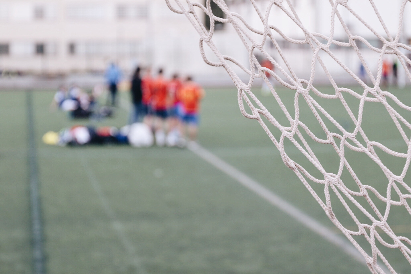 Fotografi med nærblide av nettingen i et fotballmål. I bakgrunnen ser vi fotballspillere som står på sidelinjen.