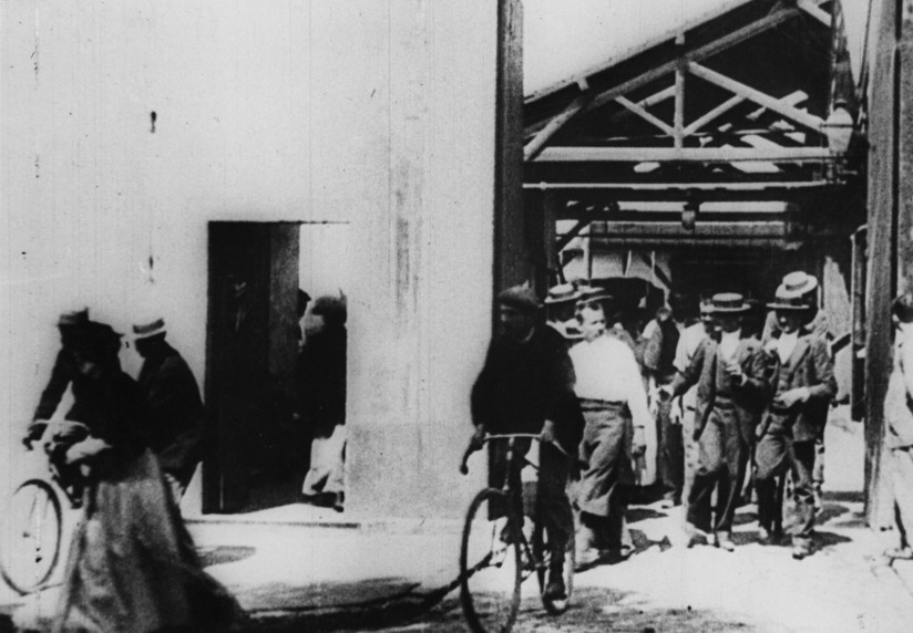 Svart-hvitt fotografi hentet fra filmen Arbeiderne forlater fabrikken som ble laget av Auguste og Louis Lumiere i 1895. Fotografiet viser arbeidere som forlater fabrikken (på sykkel og til fots) i lunsjpausen.