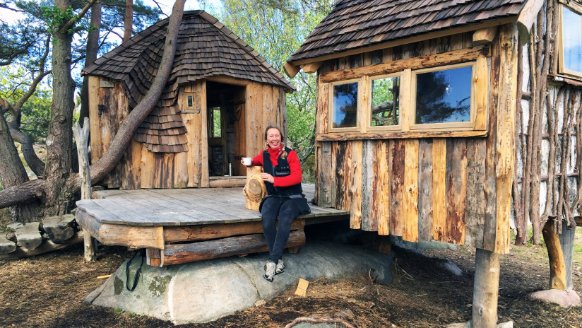 Fotografi av en kvinne som sitter på trammen utenfor en rustikk hytte. Hun holder en kopp i hånden og smiler mot fotografen.