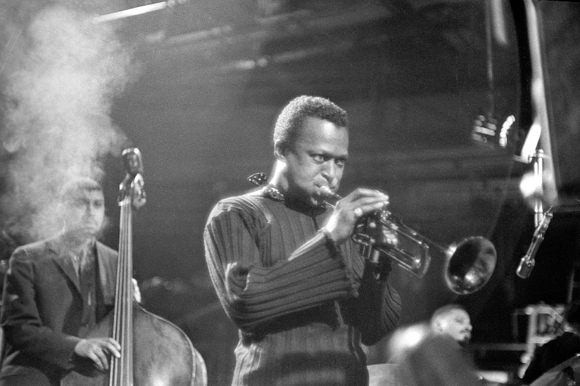 Svart-hvitt fotografi av Miles Davis (1926–1991) som spiller på en trompet i New York i 1959. Bak Davis ser vi en musiker som spiller på kontrabass samt en pianist.